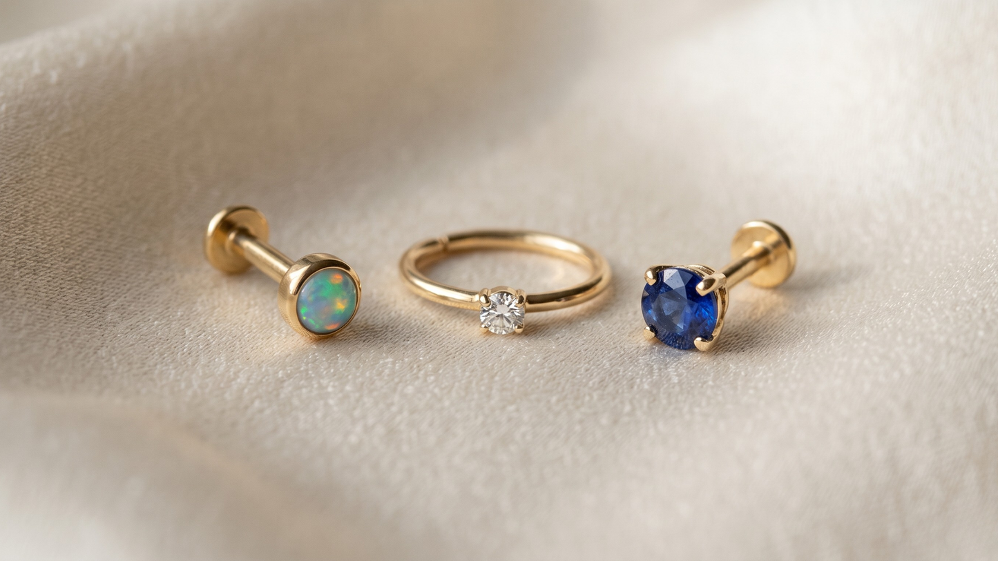 three different gemstone-set piercing pieces arranged on cream silk: a piece with a small opal (visible iridescent play of colour), a piece with a tiny natural diamond (sparkle visible), and a piece with a coloured sapphire